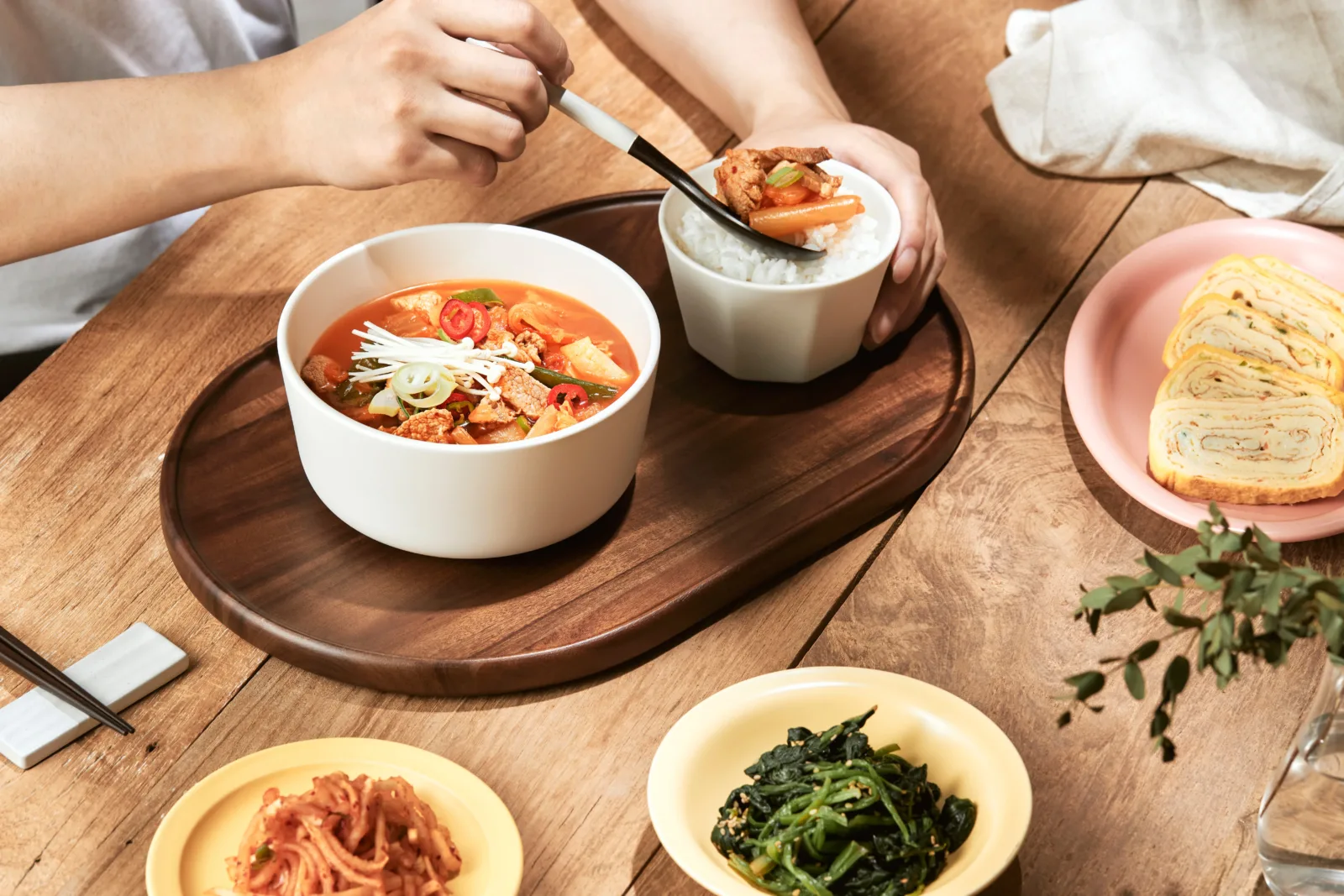나무 쟁반에 놓인 김치찌개 한 그릇과 밥, 달걀말이와 나물 반찬을 두 사람이 나눠 먹는 상